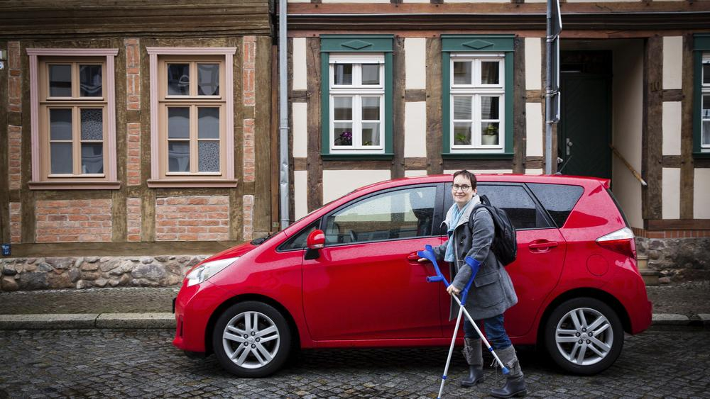 Agnes Kunze steht mit ihren Krücken vor ihrem roten Auto.