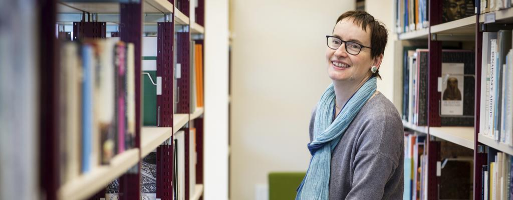 Agnes steht zwischen Bücherregalen in der Bibliothek und lächelt in die Kamera.