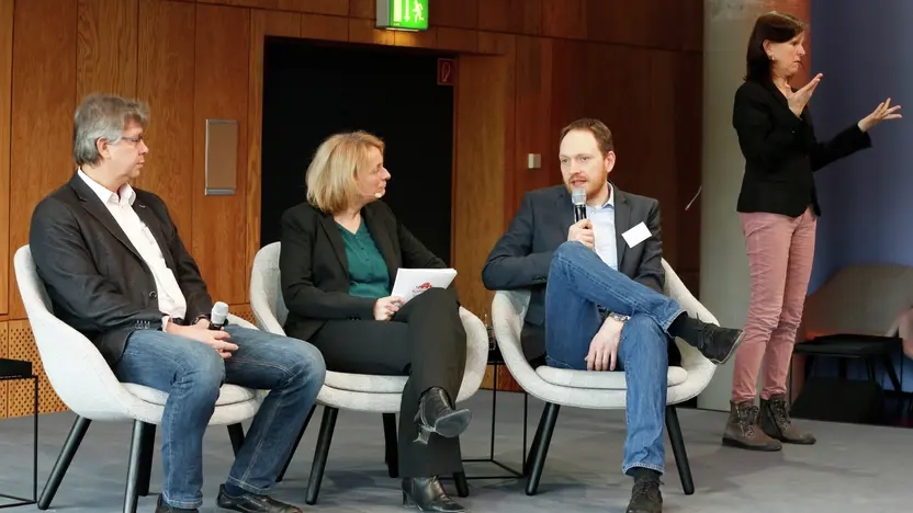 Auf einer Bühne sitzen drei Personen: Professor Dieter Katzenbach sitzt links, in der Mitte eine Moderatorin, Professor Hendrik Trescher sitzt rechts und spricht in ein Mikrofon. Rechts daneben steht eine Gebärdensprach-Dolmetscherin und übersetzt in Gebärdensprache.