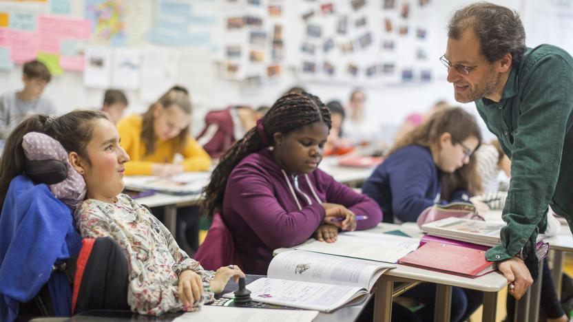 Kinder im Klassenraum an Schreibtischen. Ein Lehrer beugt sich zu einem Mädchen im Rollstuhl.