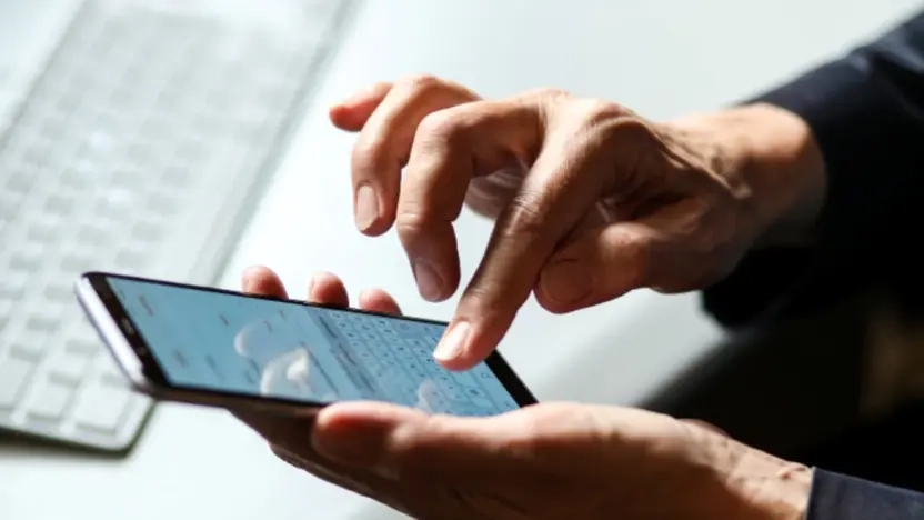 zwei haende bedienen ein smartphone. im hintergrund ist ein schreibtisch mit tastatur und computermaus zu sehen.