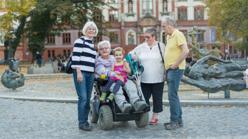 Vier Männer und Frauen, davon eine im Rollstuhl sowie ein Kleinkind vor einer Stadtkulisse.