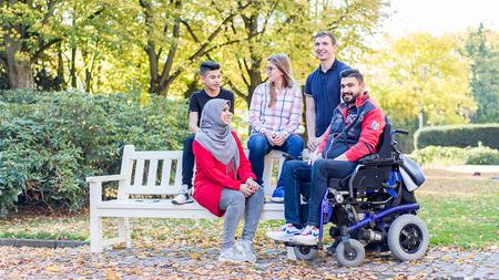 Eine Gruppe junger Menschen trifft sich an einer Bank im Rosengarten. Ein Junger Mann ist Rollstuhlfahrer.