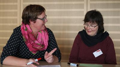 Zwei Frauen sitzen an einem Tisch. Die eine Frau erklärt der anderen Frau etwas.