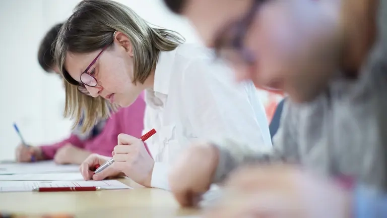 Drei Personen beugen sich über Schriftstücke, die vor ihnen liegen. Nur eine von ihnen ist scharf zuerkennen. Die anderen beiden sind unscharf oder verdeckt.