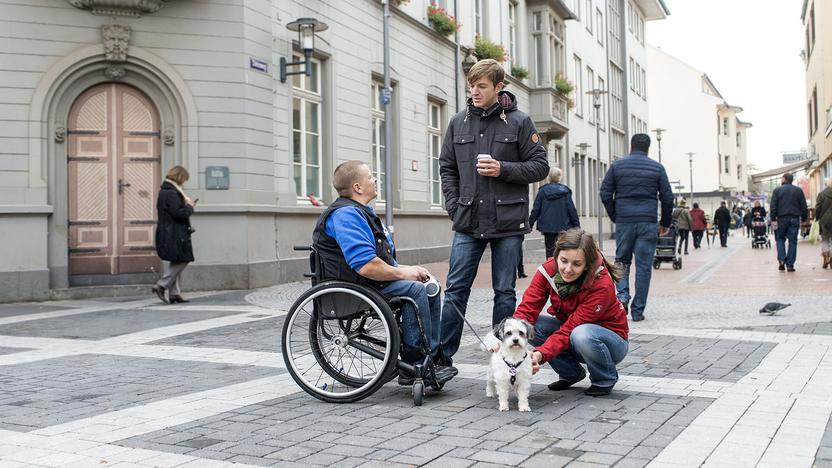 ein rollstuhlfahrer, ein junger mann und eine junge Frau mit kleinem weissen hund stehen in einer fußgängerzone