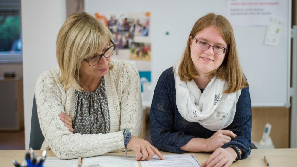 Zwei Frauen sitzen an einem Tisch und besprechen ein Dokument. Eine der Frauen sieht in die Kamera.