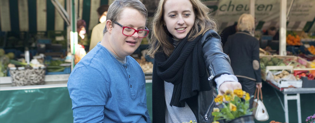 Junger Mann mit Down-Syndrom und eine junge Frau betrachten Blumen vor einem Marktstand