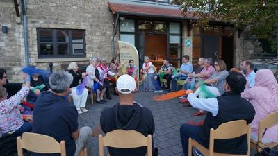 Viele Menschen sitzen im Freien in einem Stuhlkreis, ein paar von ihnen haben bunte Tücher in der Hand.