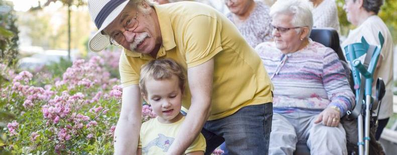 Ein älterer Mann und ein Kleinkind arbeiten im Garten, im Hintergrund arbeiten noch mehr Menschen im Garten, eine Frau sitzt im Rollstuhl