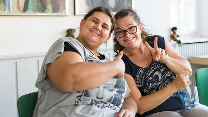 Zwei Frauen lachen in die Kamera. Sie zeigen das Victoriezeichen und Daumen Hoch mit den Händen.