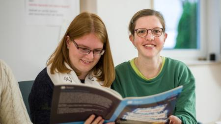 Zwei Frauen sitzen an einem Schreibtisch und halten ein Buch in den Händen, die linke Frau liest, die rechte Frau lächelt in die Kamera.