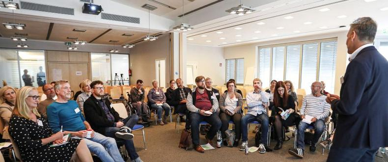 Eine Gruppe von etwa 20 Menschen hört dem Vortrag eines Mannes zu.