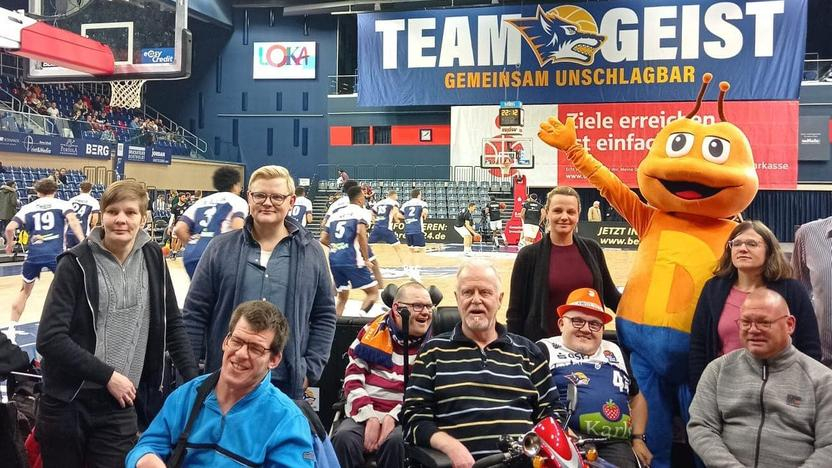 Menschen mit und ohne Behinderung stehen und sitzen vor dem Spielfeldrand der Rostock Seawolves. Im Hintergrund wärmen sich die Rostock Seawolves auf.