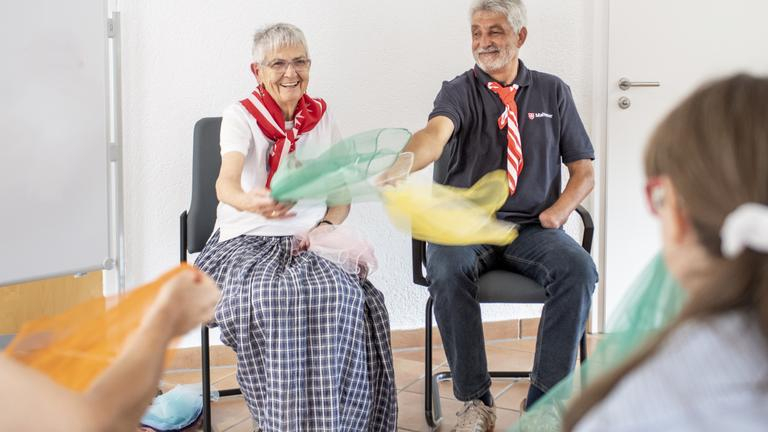 Eine ältere Frau und ein älterer Mann machen Sitztanzen mit Tüchern.