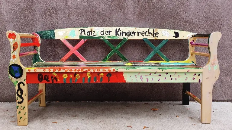 Eine bunte Holzbank auf der "Platz der Kinderrechte" steht.