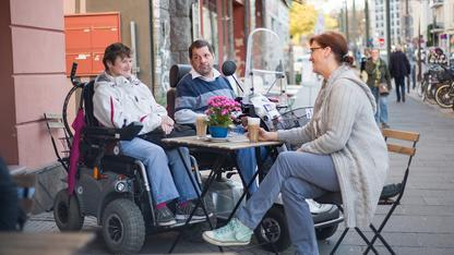 Zwei Rollstuhlfahrer und eine Fußgängerin sitzen in eienm Straßencafé und trinken Kaffee