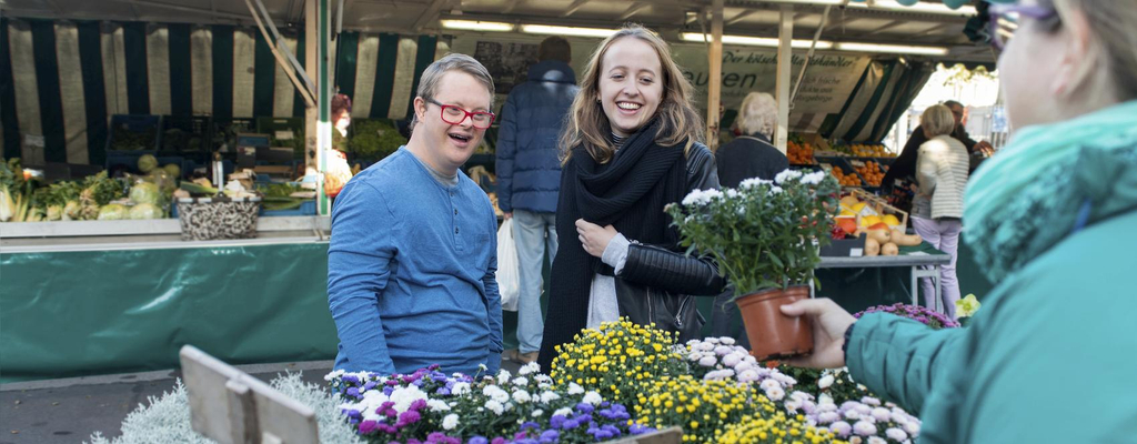 Ein junger Mann und eine junge Frau schauen sich Blumen an einem Marktstand an. Der junge Mann hat das Down-Syndrom.