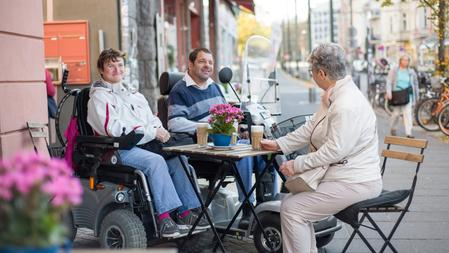 Zwei Rollstuhlfahrer*innen und eine Frau sitzen an einem Tisch im Straßencafé und unterhalten sich