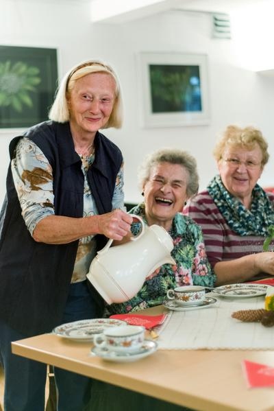 Drei ältere Frauen sitzen beziehungsweise stehen an einem Tisch und lachen in die Kamera.