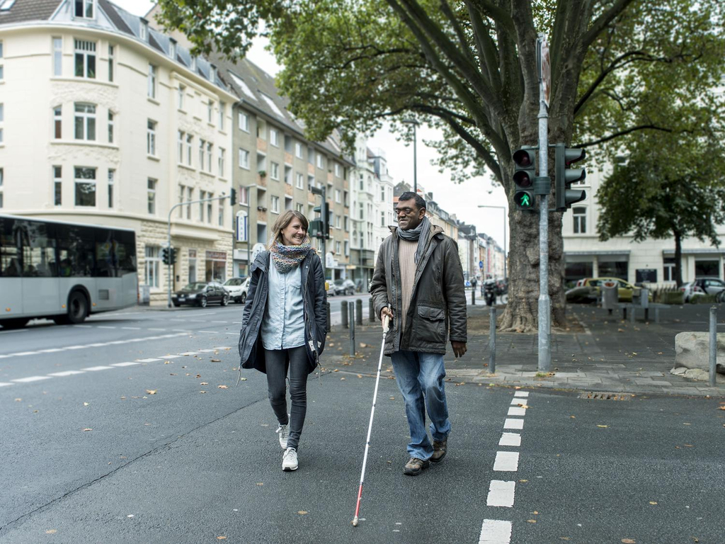Eine Frau und ein Mann mit Langstock gehen über eine Ampel.