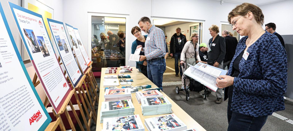 Ein Tisch voller Infobroschüren, dahinter Plakate mit Fotos und Text, davor Menschen, die durch die Broschüren blättern und die Plakate ansehen