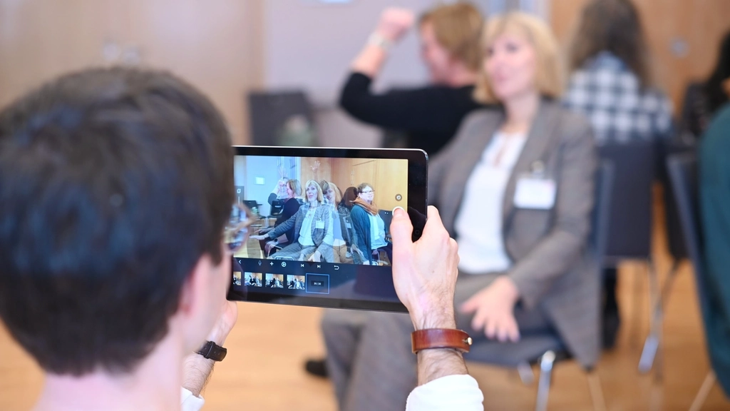 Eine Person macht mit einem Tablet Aufnahmen von Personen, die im Stuhlkreis sitzen