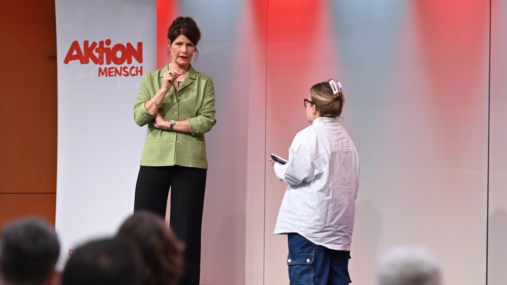 Blick auf die Bühne: Eine Frau und eine kleinwüchsige Frau im Gespräch. Rechts am Bildrand sind Gebärdensprachdolmetscherinnen.