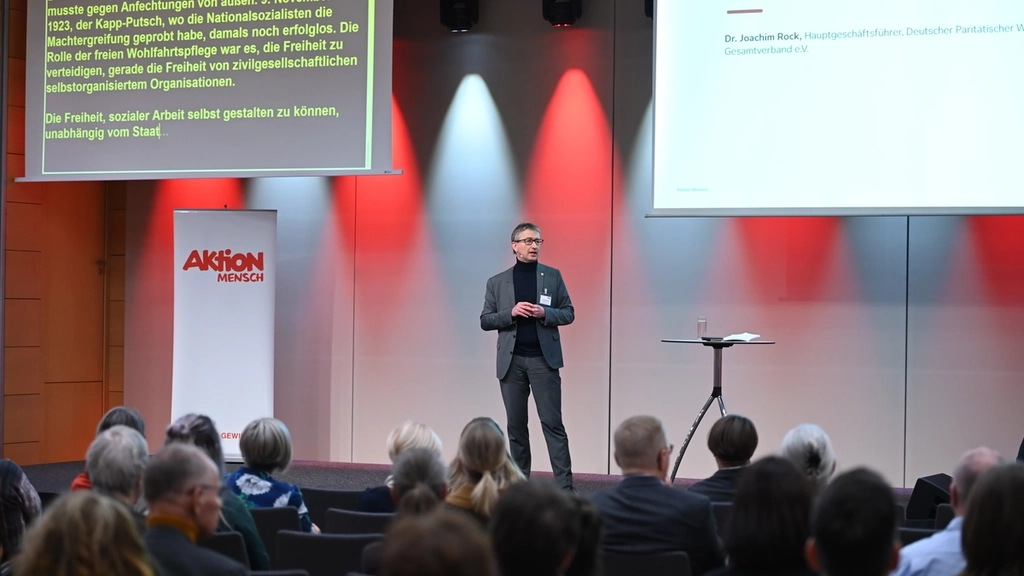 Blick auf die Bühne mit einem Referenten. Über ihm sind zwei große Bildschirme mit Schrift.