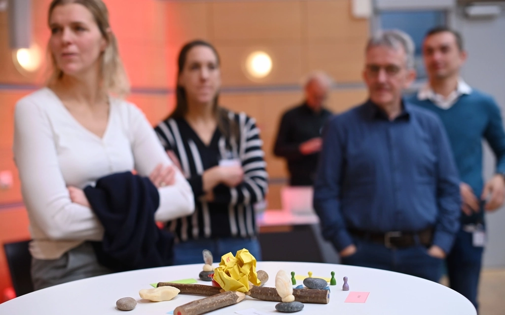 Teilnehmende eines Workshops stehen hinter einem Stehtisch, auf dem eine kleine Skultur aus verschiedenen Materialien liegt