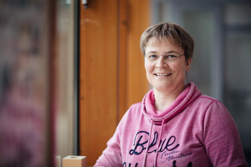 Eine Frau mit kurzen blonden Haaren und einem pinken Kapuzenpullover steht in einem Gang und lächelt in die Kamera.