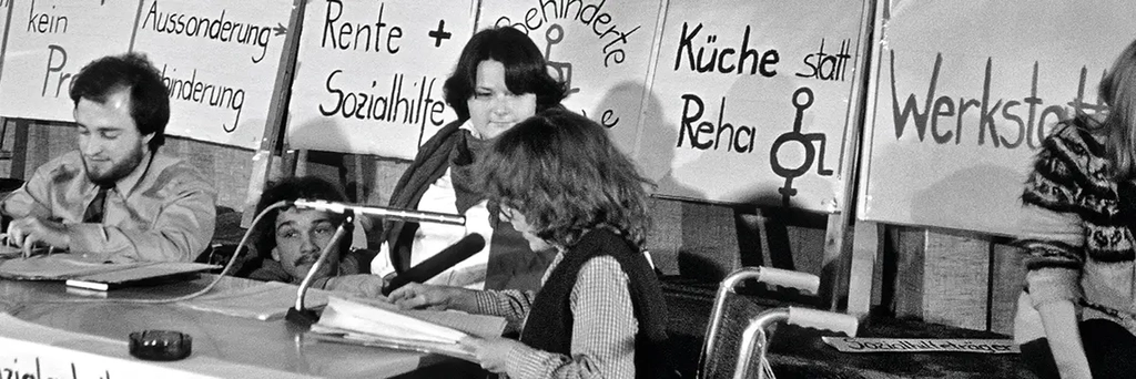 drei Menschen mit Behinderung auf einem Podium mit Mikros. Hinter ihnen stehen Transparente und Schilder mit Sprüchen zur gesellschaftlichen Situation von Menschen mit Behinderung