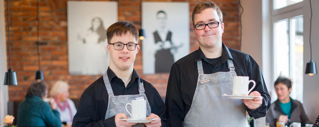 Zwei Männer mit Behinderung lächeln in die Kamera. Sie tragen Schürzen und halten Kaffeetassen in der Hand.