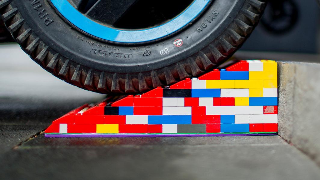 Eine bunte Legorampe liegt vor einer Stufe. Im Anschnitt ist ein Rollstuhl- oder Kinderwagen-Reifen zu sehen, der gerade über die Rampe rollt.