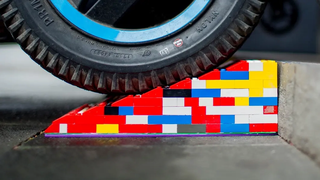 Eine bunte Legorampe liegt vor einer Stufe. Im Anschnitt ist ein Rollstuhl- oder Kinderwagen-Reifen zu sehen, der gerade über die Rampe rollt.