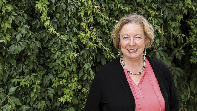 Eine Frau mit mittellangem, blonden Haar steht lachelnd vor einer Hecke.