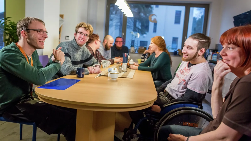 An einem Esstisch sitzen viele verschiedene Menschen mit und ohne Behinderung.