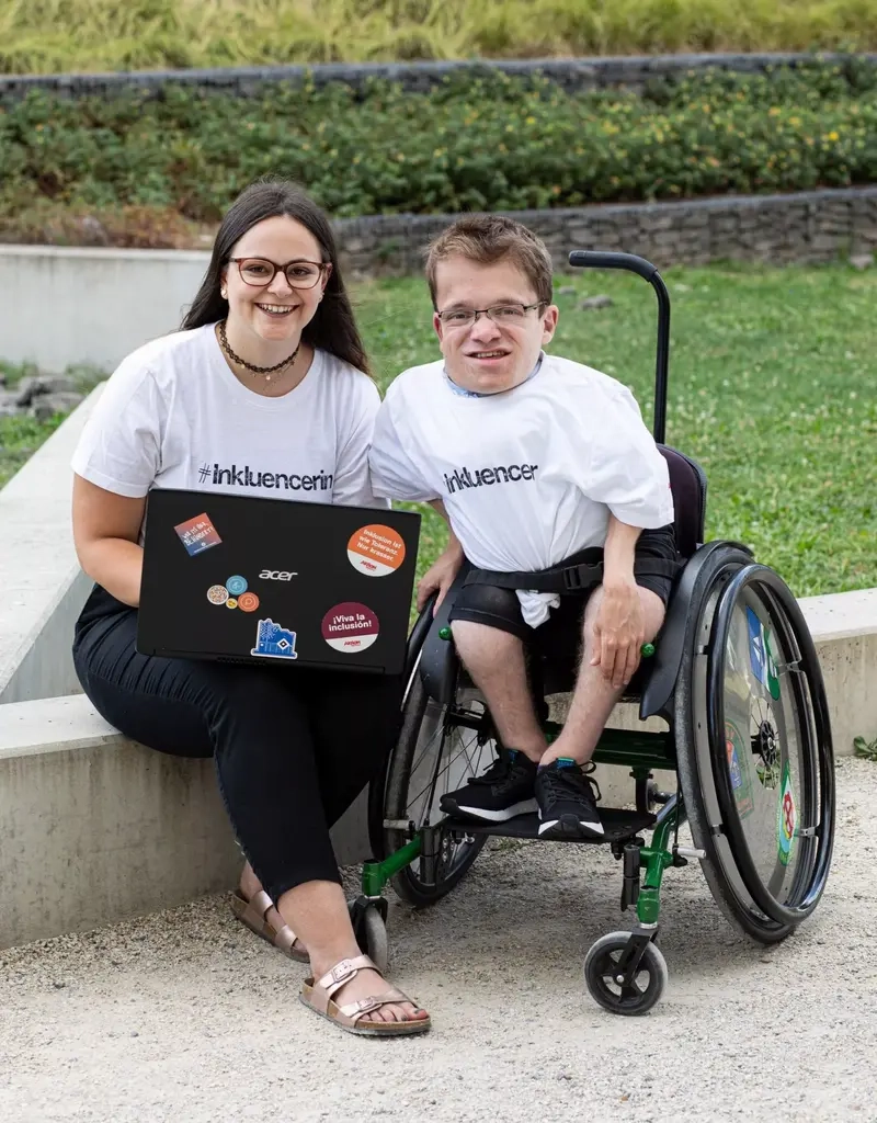 Max und Cristina sitzen in Inkluencer-T-Shirts mit einem Laptop im Freien und lächeln in die Kamera.