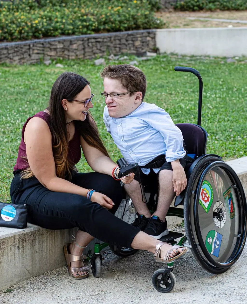 Cristina sitzt auf einer niedrigen Mauer, Max steht mit seinem Rollstuhl neben ihr. Die beiden lächeln sich verliebt an. Beide schauen sich 