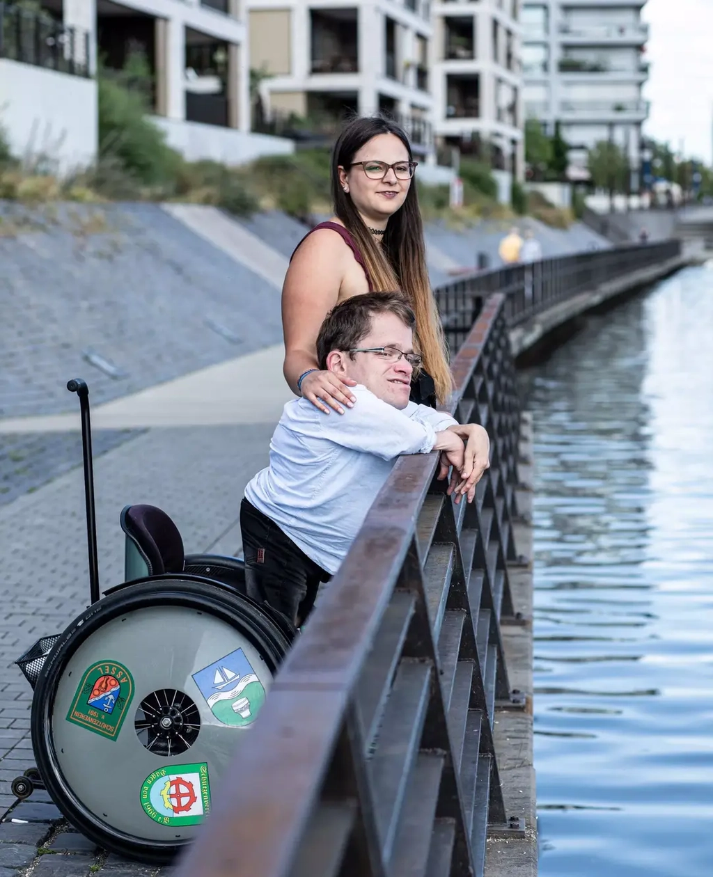 Cristina und Max stehen gemeinsam an einer Uferpromenade und schauen auf den Fluss.