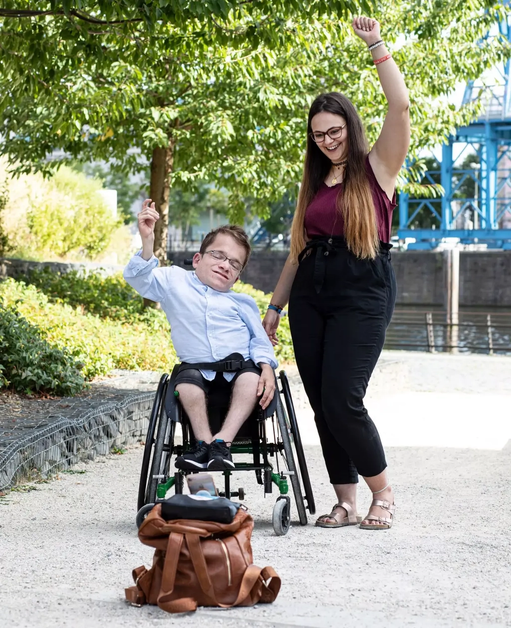 Cristina steht neben Max, der im Rollstuhl sitzt. Beide werfen die Arme in die Höhe und filmen sich dabei mit ihrem Handy für ihre Social Media-Kanäle.