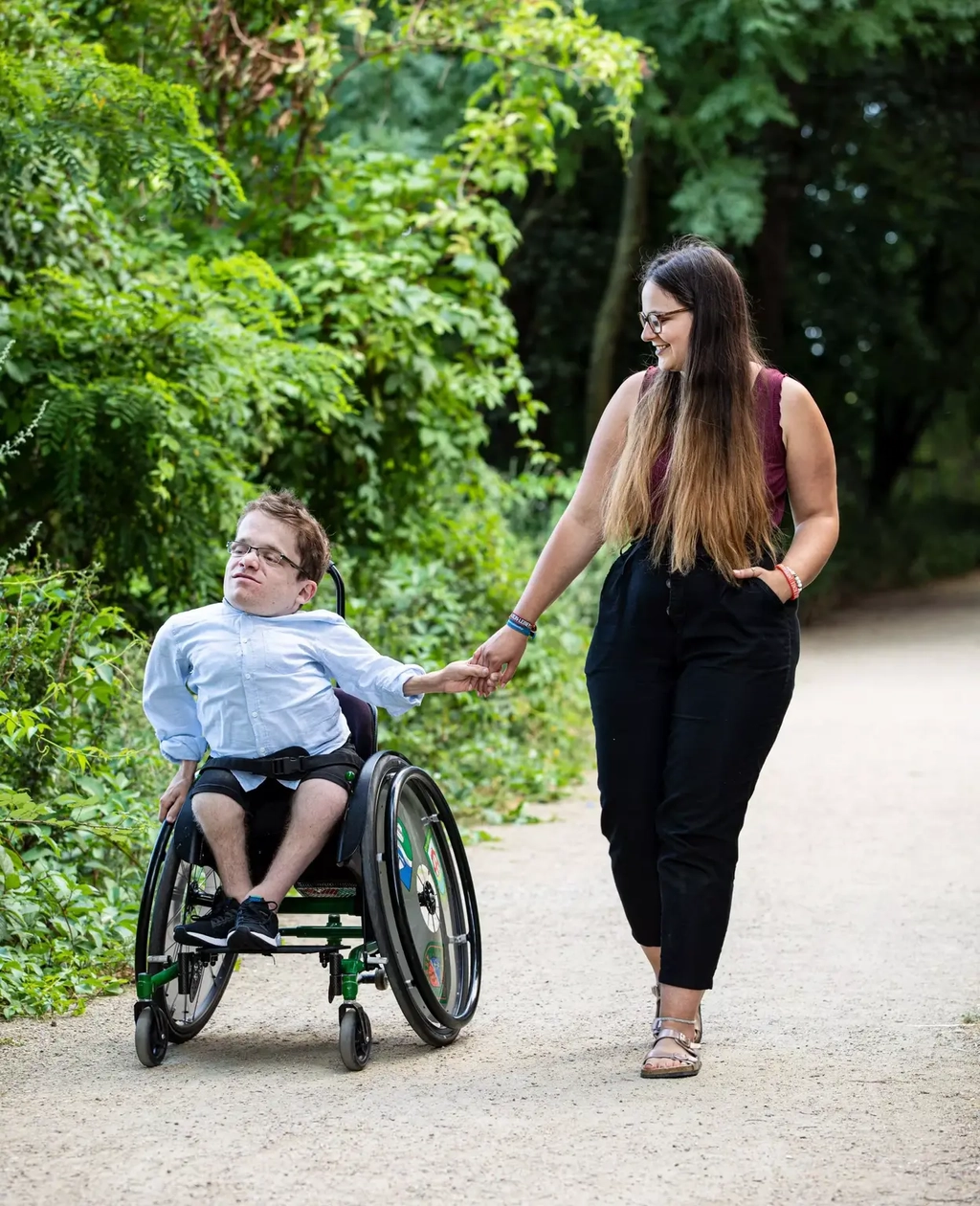 Max und Cristina machen händchenhaltend einen Spaziergang im Park. Sie läuft neben seinem Rollstuhl einen Schotterweg entlang, der von Bäumen und Büschen umgeben ist.