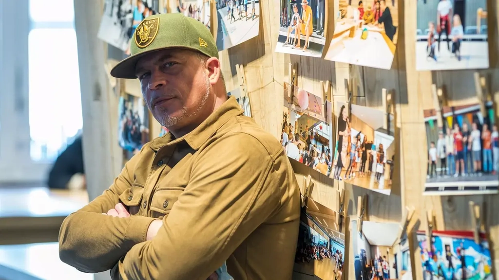 Ein Mann mit braunem Wildlederhemd und grüner Baseballmütze steht mit verschränkten Armen vor einer Fotowand. Er guckt seitlich in die Kamera.