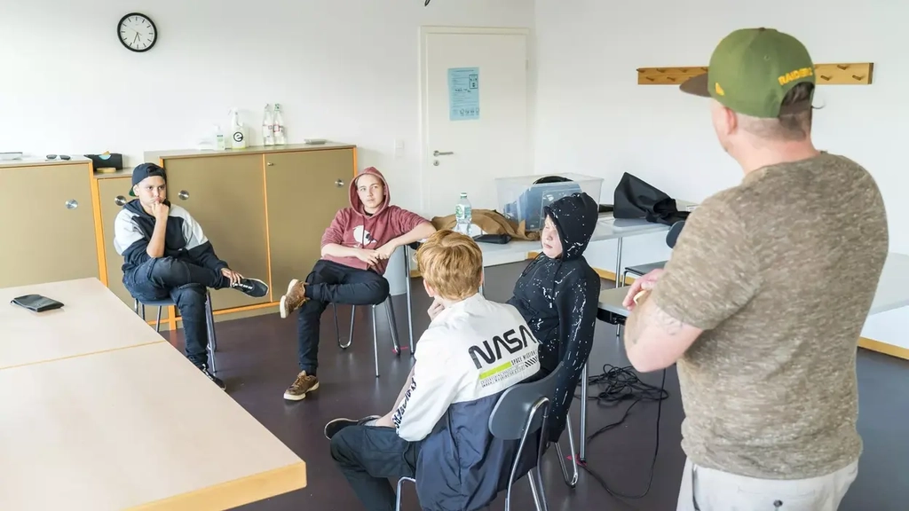 In einer Art Klassenraum sitzen vier Jugendliche auf Stühlen in der Mitte des Raums. Neben ihnen steht ein Mann mit grüner Baseball-Mütze und hört ihnen zu.