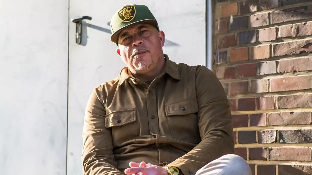 Ein Mann mit hellbraunem Hemd und heller Hose sitzt auf einer Mauer vor einer Lagerhalle. Er überkreuzt die Beine und trägt eine grüne Baseballmütze.