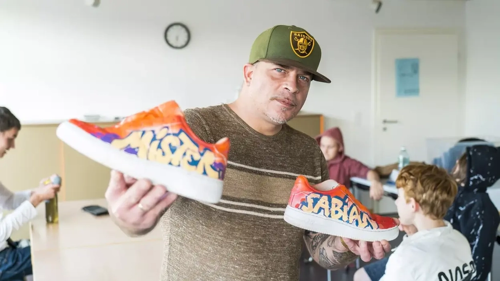Ein Mann mit grüner Baseballmütze hält in jeder Hand vor sich einen Turnschuh in die Luft. Die Schuhe sind im Graffiti-Stil bemalt.