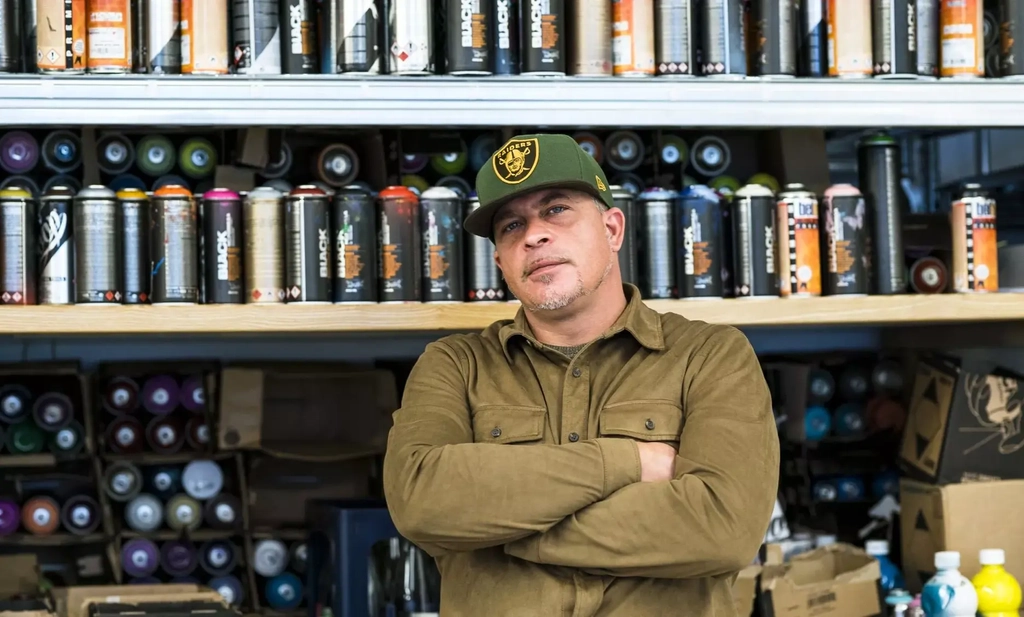 Ein Mann mit Baseballmütze und braunem Hemd steht mit verschränkten Armen vor einem Regal voller Spraydosen.