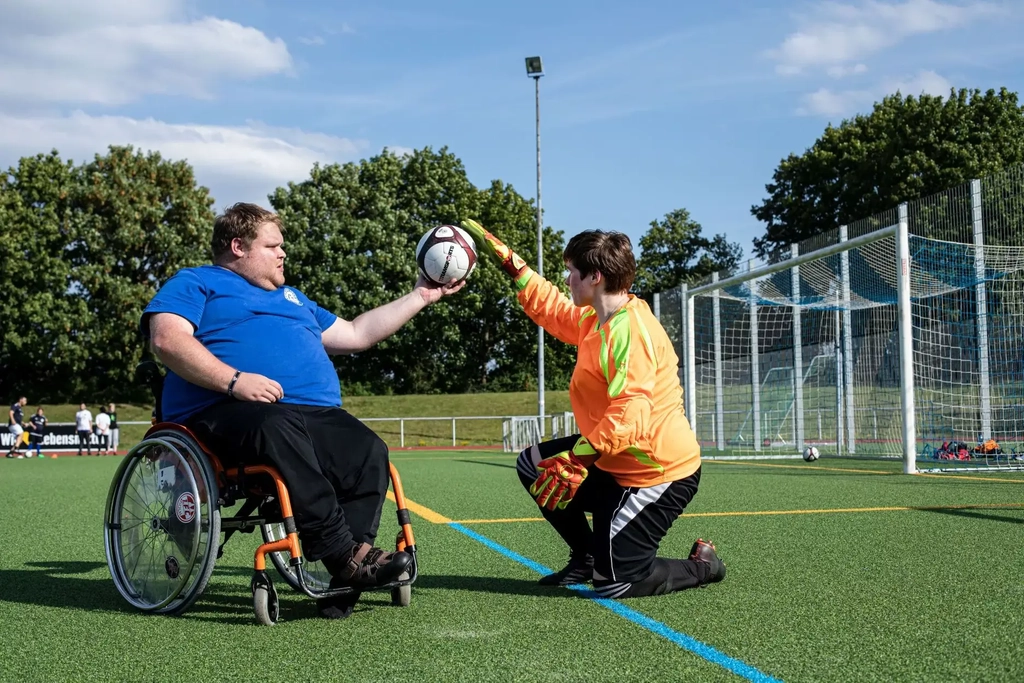 Torwarttrainer Sven sitzt im Rollstuhl und hält einen Ball in die Luft. Torwärtin Jana legt ihre Hand auf den Ball.