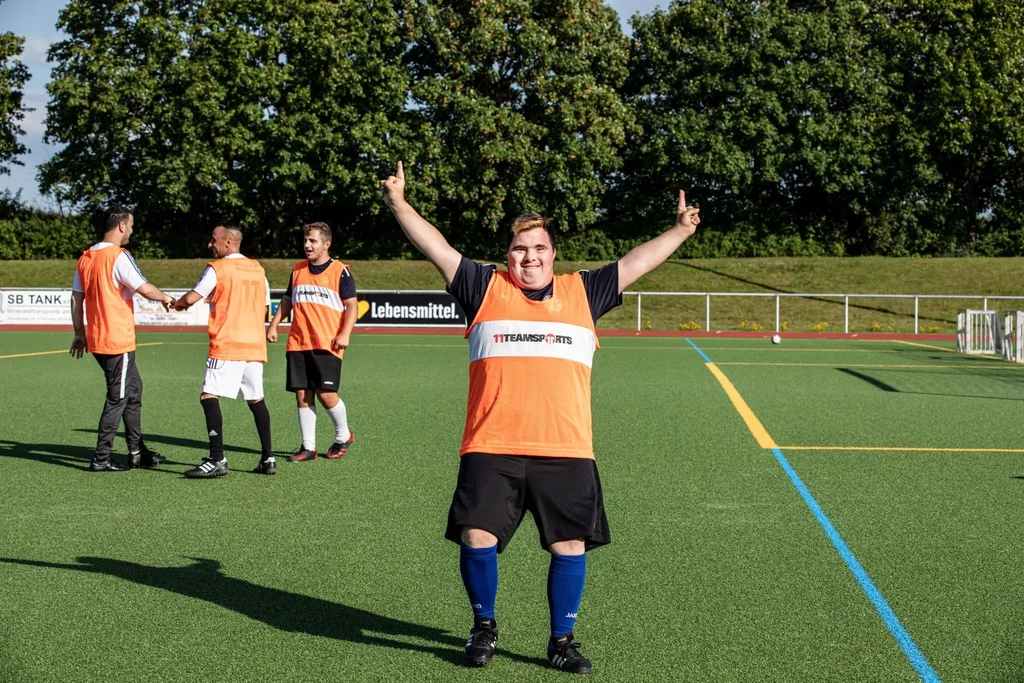 Ein Spieler des FC Germania Zündorf steht mit einem orangefarbenen Leibchen auf dem Fußballplatz und reißt siegessicher die Arme in die Luft.