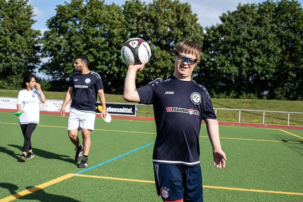 Spieler Tobias geht auf einem Fußballplatz auf die Kamera zu und hält mit der rechten Hand einen Fußball in die Luft.